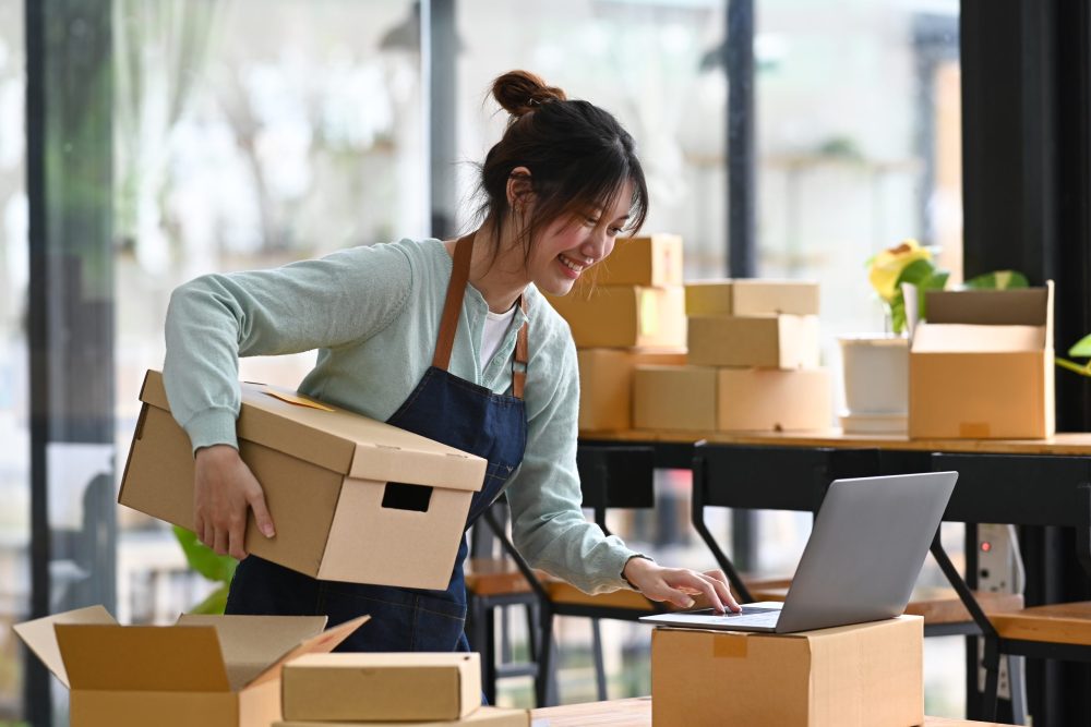 Young woman online seller holding cardboard box and using laptop for checking order on internet. Concept of online selling, e-commerce